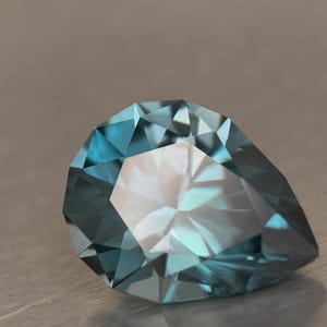 Puede incluir: Una gema facetada en forma de pera, de color turquesa. La superficie de la gema refleja la luz, mostrando sus m&uacute;ltiples &aacute;ngulos y estructura interna. La gema se encuentra sobre un fondo neutro.