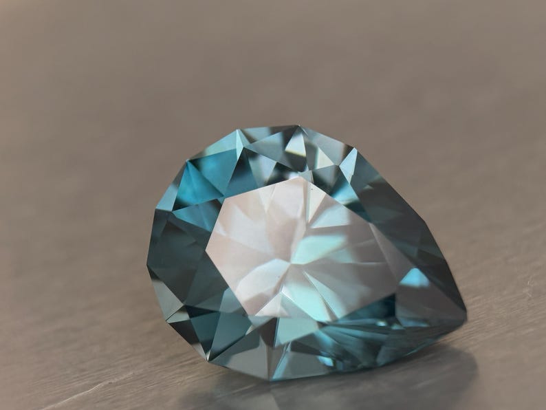 Puede incluir: Una gema facetada en forma de pera, de color turquesa. La superficie de la gema refleja la luz, mostrando sus m&uacute;ltiples &aacute;ngulos y estructura interna. La gema se encuentra sobre un fondo neutro.
