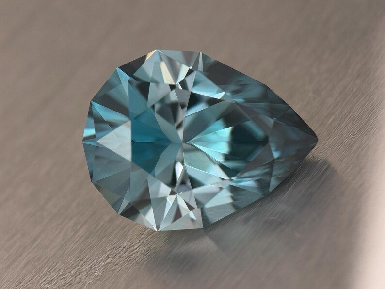 Puede incluir: Una gema facetada en forma de pera, de color azul claro. La superficie de la gema refleja la luz, mostrando sus m&uacute;ltiples facetas y su estructura interna. La gema se encuentra sobre un fondo met&aacute;lico neutro.