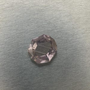 May include: A faceted, light purple gemstone with a multi-sided shape. The gem reflects light, creating a sparkling effect. The stone is set against a textured, gray surface.