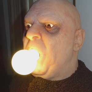 May include: A detailed, realistic sculpture of a bald head with a light bulb in its mouth. The head has a weathered appearance with visible wrinkles and dark circles under the eyes. The bulb emits a bright, warm light, creating a striking visual effect.