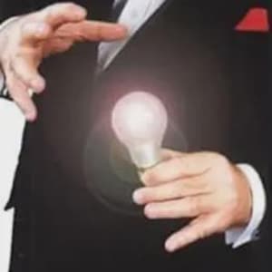 May include: A person in a black suit holds a glowing light bulb. The bulb is illuminated with a soft, pinkish light. The person's hands are positioned as if they are performing a magic trick, with one hand raised and the other holding the bulb.