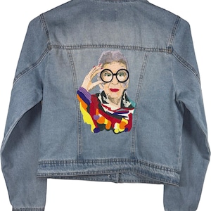 May include: Light blue denim jacket with an embroidered portrait on the back. The portrait features a woman with round glasses, red lipstick, and a colorful top. The jacket has a classic design with a collar, button closures, and pockets.