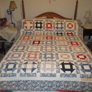 May include: A white quilt with a blue and red pattern. The quilt is made up of squares with different designs, including stars, circles, and squares. The quilt is on a bed with a wooden headboard.
