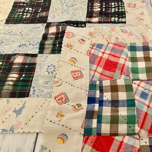 May include: A patchwork quilt made of various fabrics, including plaid, floral, and a fabric with a pattern of toy blocks. The quilt is sewn together in a grid pattern and has a white background.