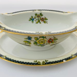 May include: A white porcelain gravy boat with a gold rim and floral pattern. The boat is on a matching saucer with a gold rim and floral pattern.