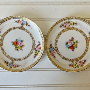 May include: Two white porcelain plates with gold rims and floral designs. Each plate features a central bouquet of colorful flowers and a decorative gold border with floral accents. The plates are set against a white background.