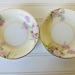 May include: Two small, white ceramic bowls with a gold rim. The bowls have a floral design with pink flowers and green leaves on a cream background.