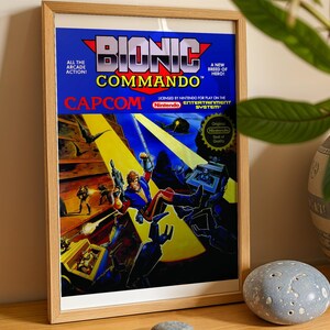 May include: A framed print of the Bionic Commando video game cover art for the Nintendo Entertainment System. The artwork features a character with a bionic arm, a blue and yellow color scheme, and the text Bionic Commando, Capcom, and Nintendo.