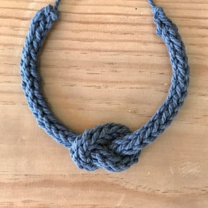 以下が含まれることがあります： 中央に装飾的な結び目がある、手作りの青いロープネックレス。 ネックレスは編みロープで作られており、シンプルで航海をテーマにしたデザインです。 明るい色の木製の表面に表示されています。