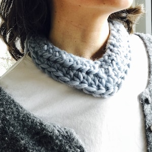 May include: A light blue knitted neck warmer or cowl, worn by a person over a white t-shirt and a gray cardigan.