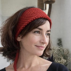 Könnte beinhalten: Ein gestricktes Stirnband in einem satten, warmen Rotton. Das breite Stirnband bedeckt die Stirn, mit einem passenden Strickband. Die Frau im Bild trägt ein schwarzes Oberteil.