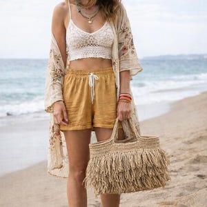 Könnte beinhalten: Ein Strand-Outfit mit Strohhut, weißem Häkeltop, senffarbenen Shorts und einer Fransen-Tragetasche. Ein Blumenkimono und mehrschichtige Halsketten vervollständigen den Look. Die Szene spielt sich an einem Sandstrand mit dem Meer im Hintergrund ab.