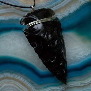 May include: A black obsidian arrowhead pendant with silver wire wrapping, displayed on a blue and white agate background.