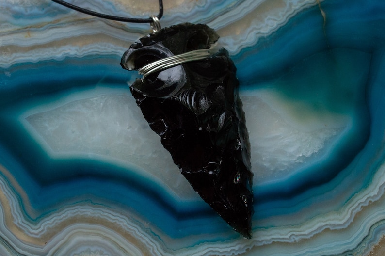 May include: A black obsidian arrowhead pendant with silver wire wrapping, displayed on a blue and white agate background.