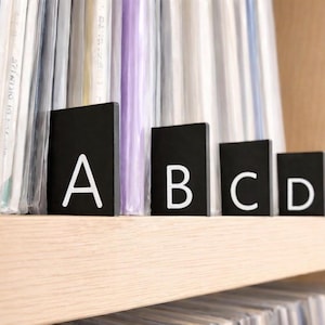 May include: A set of four rectangular black dividers with white letters A, B, C, and D printed on the front. These dividers are positioned upright to organize a collection of items stored in clear plastic sleeves on a wooden shelf.