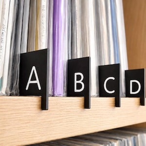 Può includere: Divisori per dischi neri con lettere bianche A, B, C e D, posizionati su una mensola di legno. Vengono utilizzati per organizzare una collezione di dischi in vinile.