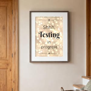Op de afbeelding: Een ingelijste print met de woorden "Shhh, Testing in progress" in een schreefletter, tegen een achtergrond van zachte, onscherpe lichten. De print wordt weergegeven op een beige muur, in de buurt van een houten deur en een rieten mand.