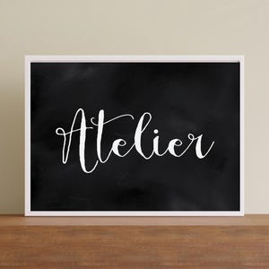 Op de afbeelding: Een rechthoekig wandkunstwerk met een zwarte achtergrond met een subtiele, bewolkte textuur en het woord Atelier geschreven in wit, elegant cursief schrift. Het kunstwerk is ingelijst in een dunne, witte lijst voor een contrastrijk beeld.