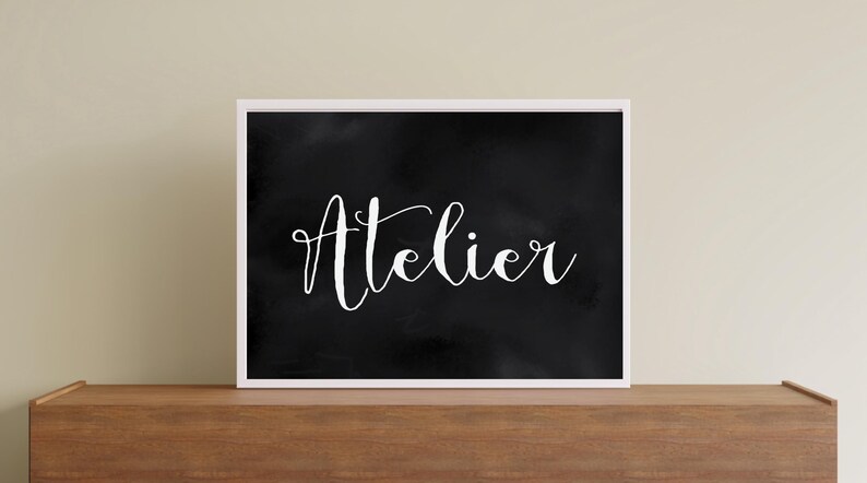 Op de afbeelding: Een rechthoekig wandkunstwerk met een zwarte achtergrond met een subtiele, bewolkte textuur en het woord Atelier geschreven in wit, elegant cursief schrift. Het kunstwerk is ingelijst in een dunne, witte lijst voor een contrastrijk beeld.