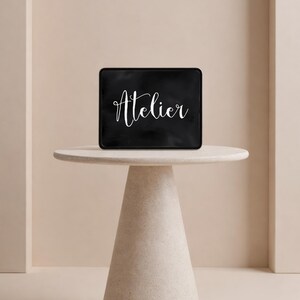 Op de afbeelding: Een rechthoekig zwart bord met afgeronde hoeken waarop het woord Atelier in witte cursieve letters staat. Het bord staat op een lichtgekleurde ronde tafel met een conische voet, gecentreerd tegen een muur in neutrale tinten.