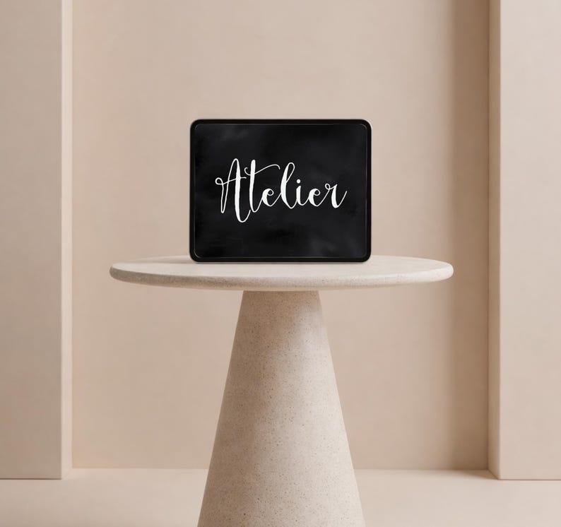 Op de afbeelding: Een rechthoekig zwart bord met afgeronde hoeken waarop het woord Atelier in witte cursieve letters staat. Het bord staat op een lichtgekleurde ronde tafel met een conische voet, gecentreerd tegen een muur in neutrale tinten.