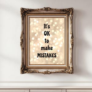 May include: A framed print featuring the message "It's OK to make MISTAKES" in black lettering. The background has a bokeh effect of warm, blurred lights. The frame is gold-coloured and elaborately decorated.