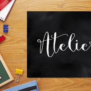 Op de afbeelding: Een rechthoekig zwart bord met het woord Atelier in witte cursieve letters. Het bord is omringd door kantoorbenodigdheden, waaronder een notitieboek, kleurpotloden, paperclips, een klein schoolbord met krijtjes en een blauwe etui.