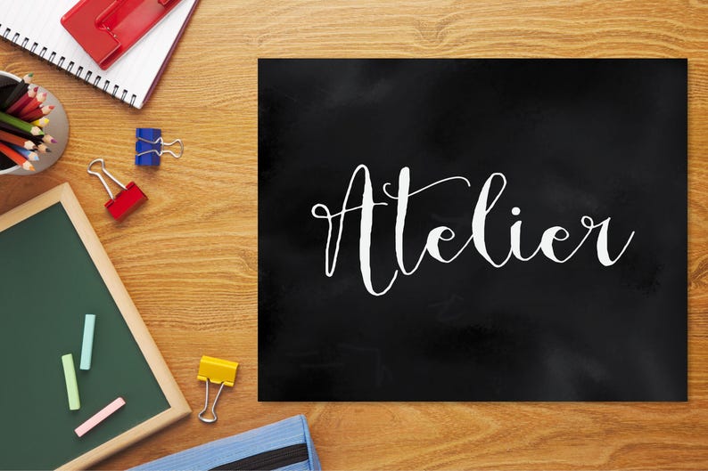 Op de afbeelding: Een rechthoekig zwart bord met het woord Atelier in witte cursieve letters. Het bord is omringd door kantoorbenodigdheden, waaronder een notitieboek, kleurpotloden, paperclips, een klein schoolbord met krijtjes en een blauwe etui.