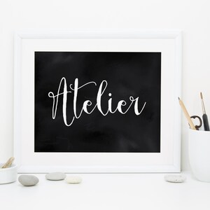 Op de afbeelding: Een wit ingelijste print met het woord Atelier in witte cursieve letters op een zwarte achtergrond. Naast de lijst staat een witte mok met verschillende dunne penselen en een klein wit schaaltje met enkele gladde grijze en witte stenen.