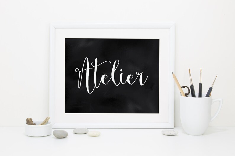 Op de afbeelding: Een wit ingelijste print met het woord Atelier in witte cursieve letters op een zwarte achtergrond. Naast de lijst staat een witte mok met verschillende dunne penselen en een klein wit schaaltje met enkele gladde grijze en witte stenen.