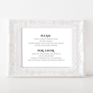 May include: A white framed sign with ornate detailing. The sign has black text that reads "PLEASE ONLY TOILET PAPER GOES IN THE TOILET EVERYTHING ELSE GOES IN THE TRASH (EVEN FLUSHABLE WIPES) POR FAVOR SOLO SE TIRA PAPEL HIGIENICO EN EL INODORO TODO LO DEMÁS VA A LA BASURA (INCLUSO LAS TOALLITAS DESECHABLES)".