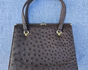 May include: A dark brown handbag with a textured ostrich-style finish. The bag features a trapezoidal shape, two top handles, and a gold-tone metal clasp closure at the center. The hardware connecting the handles to the bag is also gold-tone.