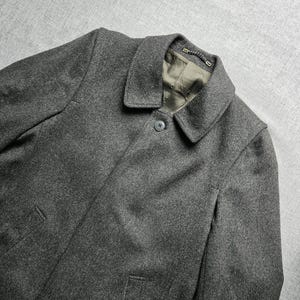 Könnte beinhalten: Ein anthrazitfarbener Mantel mit klassischem Kragen, einem einzelnen sichtbaren Knopfverschluss am Hals und zwei vertikalen Eingrifftaschen. Das Kleidungsstück ist mit einem olivgrünen Stoff gefüttert und hat eine kleine Aufhängeschlaufe.