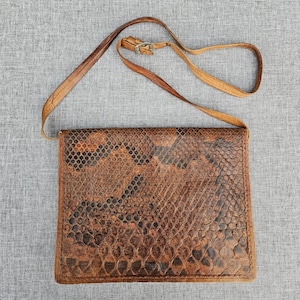 Op de afbeelding: Een rechthoekige schoudertas met een bruin en zwart slangenleerpatroon. De tas heeft een overslagsluiting en een dunne, verstelbare bruine leren schouderriem met een kleine metalen gesp voor het aanpassen van de lengte.