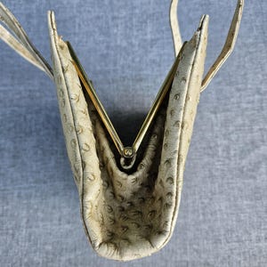 May include: A cream-colored handbag with a textured ostrich-skin pattern and a gold-tone metal frame closure. The bag features a structured design with a top handle and is shown from an overhead perspective, highlighting the open V-shaped frame.