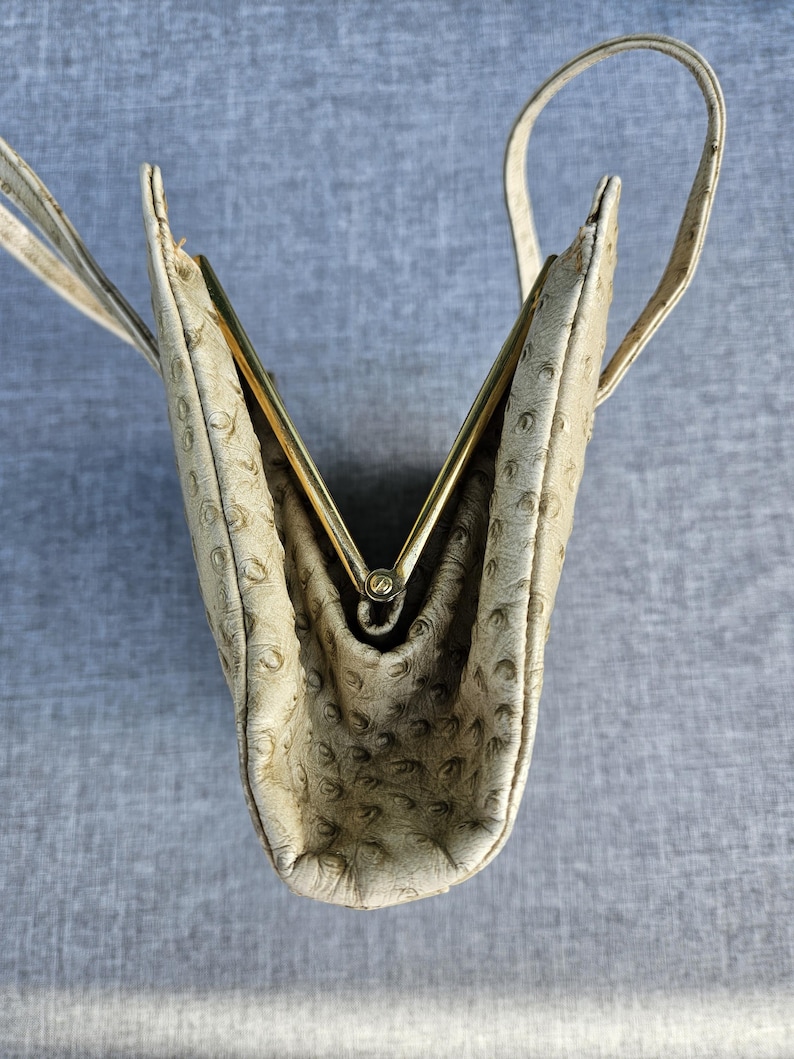 May include: A cream-colored handbag with a textured ostrich-skin pattern and a gold-tone metal frame closure. The bag features a structured design with a top handle and is shown from an overhead perspective, highlighting the open V-shaped frame.