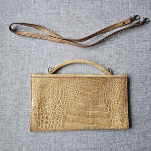 May include: A tan, rectangular handbag with a crocodile skin pattern. It has a curved handle and a detachable shoulder strap. The bag is made of a textured material and is set against a grey background.