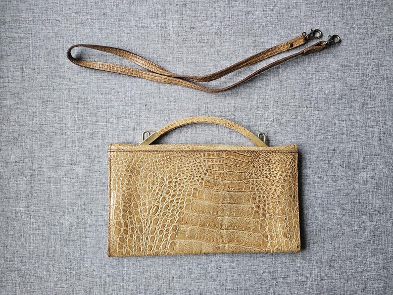 May include: A tan, rectangular handbag with a crocodile skin pattern. It has a curved handle and a detachable shoulder strap. The bag is made of a textured material and is set against a grey background.
