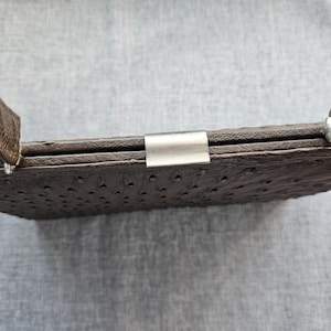 May include: A dark brown handbag with a textured, dotted surface pattern resembling ostrich leather. The bag features a rectangular frame, a silver-toned metal clasp closure at the top center, and two matching brown handles attached to the sides.