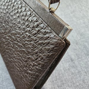May include: A dark brown handbag with a textured, bumpy surface resembling ostrich leather. The bag features a structured frame, a single top handle attached with silver-toned metal hardware, and a rectangular shape with visible stitching along the edges.