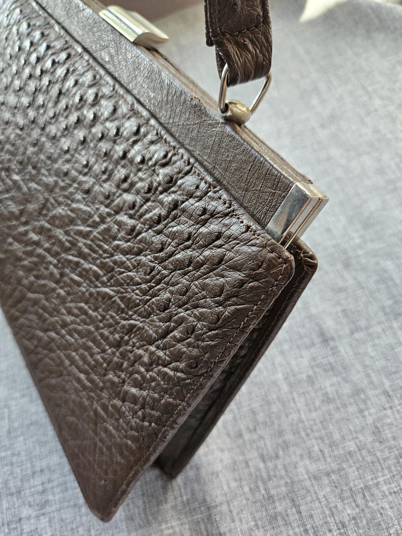 May include: A dark brown handbag with a textured, bumpy surface resembling ostrich leather. The bag features a structured frame, a single top handle attached with silver-toned metal hardware, and a rectangular shape with visible stitching along the edges.