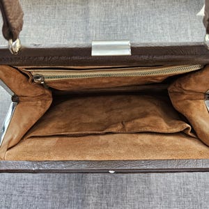 May include: A brown handbag with a textured exterior and a silver-toned metal frame closure. The interior is lined with a tan suede-like material and features a single zippered pocket along the top edge and an open slip pocket on the main compartment wall.
