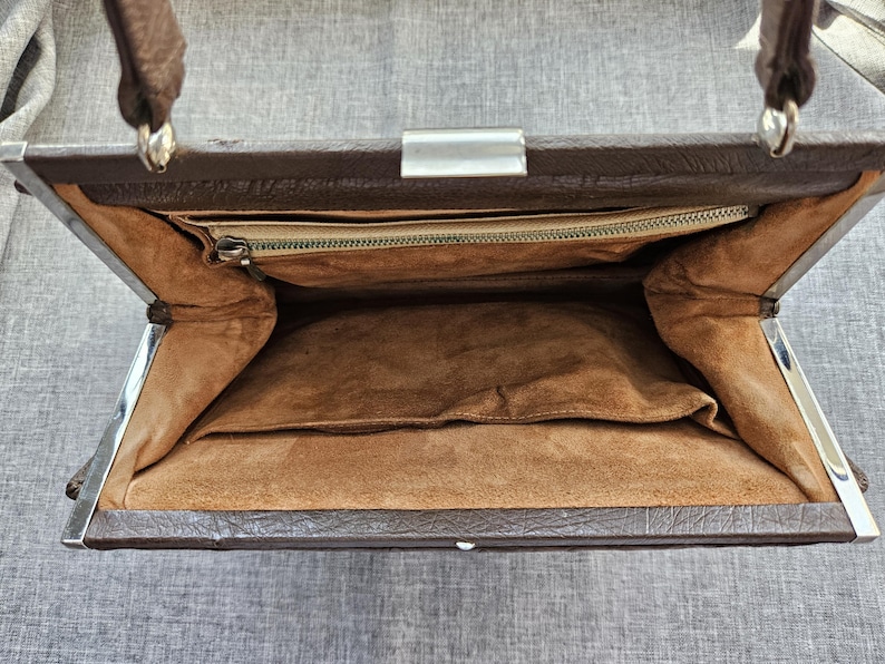 May include: A brown handbag with a textured exterior and a silver-toned metal frame closure. The interior is lined with a tan suede-like material and features a single zippered pocket along the top edge and an open slip pocket on the main compartment wall.