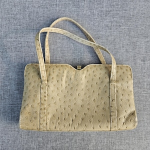 May include: A beige handbag with a textured ostrich-skin pattern, featuring two thin top handles, a rectangular body with a slightly curved top edge, and a small gold-toned metal clasp centered at the top opening of the bag.