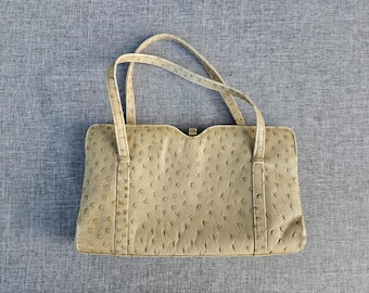 May include: A beige handbag with a textured ostrich-skin pattern, featuring two thin top handles, a rectangular body with a slightly curved top edge, and a small gold-toned metal clasp centered at the top opening of the bag.