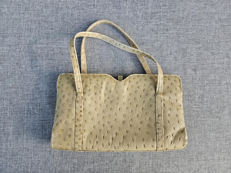 May include: A beige handbag with a textured ostrich-skin pattern, featuring two thin top handles, a rectangular body with a slightly curved top edge, and a small gold-toned metal clasp centered at the top opening of the bag.