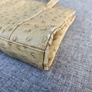 May include: A beige clutch with a textured ostrich-style pattern featuring raised circular bumps across the surface. The rectangular accessory has stitched edges and a folded construction, showing a light tan color with darker shading along the seams.