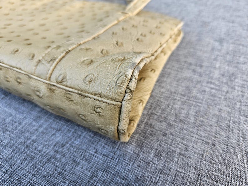 May include: A beige clutch with a textured ostrich-style pattern featuring raised circular bumps across the surface. The rectangular accessory has stitched edges and a folded construction, showing a light tan color with darker shading along the seams.