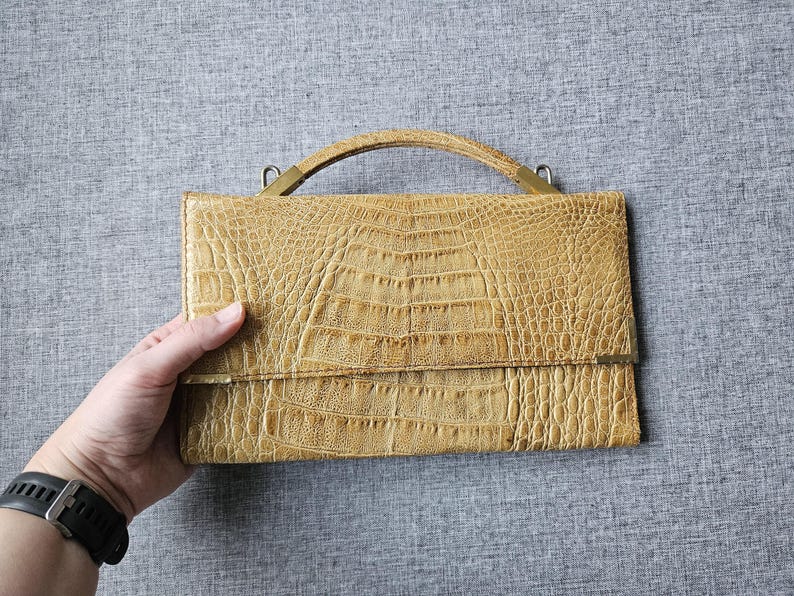May include: A tan, rectangular handbag with a textured, crocodile-like pattern. The bag has a small handle and metal hardware. The bag is held by a person.